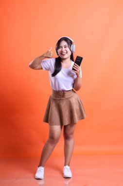 Tüm vücuduyla neşeli Asyalı bir kadın cep telefonu aletini kafasının yanında tutuyor. turuncu arkaplanda izole