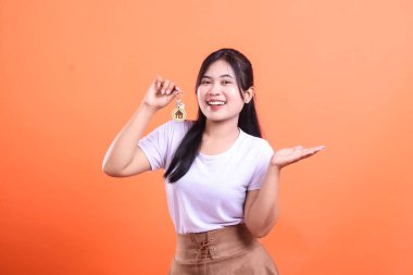 Gülümseyen genç bir kadın, bir elinde ev ikonu, bir elinde de sunum jesti olan bir anahtar tutuyor. turuncu arkaplanda izole