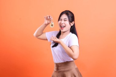 Gülümseyen genç bir kadın yeni evinin anahtarlarını tutuyor, turuncu arka planda izole edilmiş yeni evi için mutluluk ve heyecan sergiliyor.