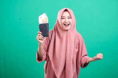 Sevinçli bir Müslüman kadın, yeşil arka planda izole edilmiş bir şekilde avro para birimiyle dolu bir cüzdan taşıyor, refah ve başarı taşıyor. Bu resim onun finansal zaferini gösteriyor..