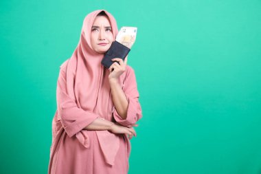 Elinde avro banknotları olan tesettürlü Müslüman bir kadın, yüzünde bir endişe ifadesi var, muhtemelen borçları düşünüyor, yeşil bir arka planda izole edilmiş..