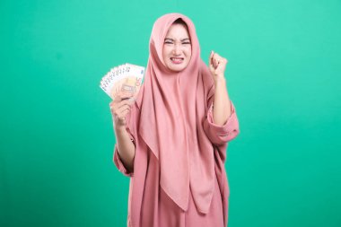 Bir tomar avro banknotları gösteren tesettürlü kızgın Müslüman kadın, mali zorluk ve hüsranı resmediyor; yeşil bir arka planda izole edilmiş.