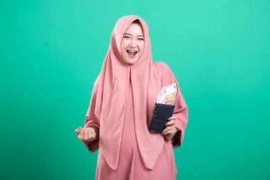 Genç Müslüman kadın, yeşil bir arka planda izole edilmiş yatırımında mali başarı ve refahı simgeleyen bir cüzdan dolusu avroyla neşesini dile getiriyor..