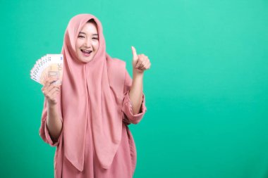 Neşeli Müslüman bir kadın bir sürü para gösteriyor ve yeşil bir arka planda izole edilmiş mutlu bir ifadeyle baş parmak işareti yapıyor. Finansal başarı ve zenginliği temsil eden.