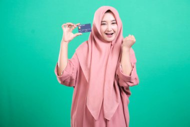 Çevrimiçi ödeme kolaylığını ve yeşil arka planda izole edilmiş modern bankacılığı temsil eden, kredi kartını gösteren mutlu bir Müslüman kadın. Finansal güvenlik ve yetkilendirmeyi somutlaştırıyor, pozitif bir hava yaratıyor..