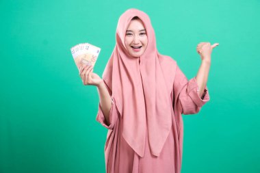 Başarılı Müslüman kadın, elinde para tutarak seviniyor ve yeşil bir arkaplanda izole bir şekilde refahını sergiliyor. Maddi bağımsızlığı ve başarıyı simgeliyor..
