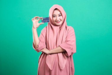 Bir tesettür takan genç Müslüman bir kadının portresi, neşeli bir şekilde bir banka kartı sunuyor, modern finans için bir kapı, yeşil arka planda izole edilmiş, para yönetimi ve yaşam tarzına modern bir yaklaşımı temsil ediyor..