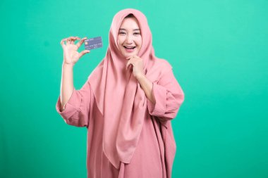 Başörtüsü içinde gülümseyen ve kredi kartı gösteren genç müslüman bir kadın, yeşil arka planda izole edilmiş, finansal erişimi ve modern kolaylığı simgeliyor. Günlük yaşam için güvenli işlemlerin gücünü gözler önüne serer