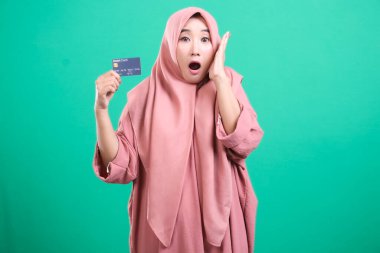 Heyecanlı müslüman kadın kredi kartını göstererek sürpriz yapıyor, yeşil bir geçmişe sahip, finansal kazancı vurguluyor ya da inanılmaz bir teklifi vurguluyor, şaşırmış hissediyor.