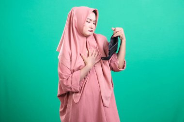 Başörtüsü içindeki endişeli Müslüman kadın üzgün bir ifadeyle boş cüzdanına bakıyor. Yeşil arka planda izole edilmiş mali kriz yüzünden stresli..