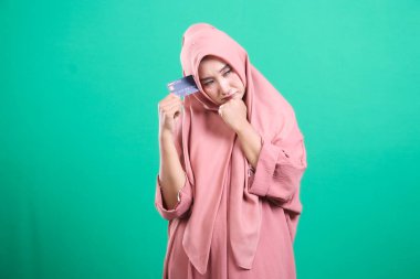 Başörtüsü takan düşünceli genç bir müslüman kadın, yeşil bir arka planda izole edilmiş mali zorluklar ve borç yönetimi ile ilgili endişelerini dile getiren bir kredi kartı tutuyor..