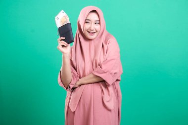 Para dolu bir cüzdan taşıyan tesettürlü neşeli genç bir kadın, mali özgürlüğü ve bolluğu sembolize ediyor, yeşil bir arka planda izole edilmiş, refah ve büyümeyi simgeliyor. Görüntü, pozitifliği ve zenginliği iletir..