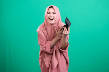 Başörtüsü içindeki genç bir kadın cüzdanını boş bulduğu için büyük ihtimalle maddi zorluklar ve belirsizliği yansıtan, yeşil bir arka planda izole edilmiş bir şekilde derin bir üzüntü yaşıyor..