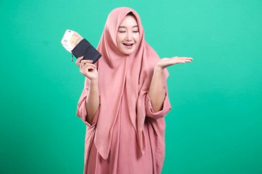 Işıl ışıl gülümseyen Asyalı güzel Müslüman kadın yeşil arka planda izole edilmiş cüzdanındaki parayı gururla sunar. Açık bir el hareketi sergiliyor. Mali planlama ve refah.