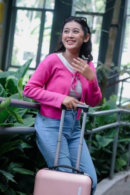 Gülümseyen genç kadın açık pembe bavulunu tutuyor, yaklaşan maceraya hazır, canlı hırkası renk katıyor, ve seyahat etmekten gerçekten mutlu görünüyor.