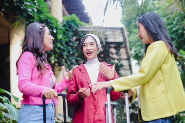 Üç gülümseyen genç kadın canlı sohbete dalarken görülüyor, bavulları bir tatil köyüne varıldığını gösteriyor. Canlı kıyafetler ve yeşillik neşeli bir tatil enerjisi hissi uyandırıyor..