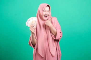 Avro banknotlarını elinde tutan heyecanlı Müslüman kadın, yeşil bir ortamda neşe ve finansal güvenlik saçıyor, yeşil bir arka planda büyüleyici bir zenginlik ve başarı görüntüsü yaratıyor,.