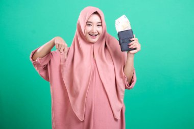 Elinde para dolu bir cüzdan olan neşeli Müslüman kadın yeşil bir arka planda izole edilmiş, finansal başarısını ve servetini sunuyor..
