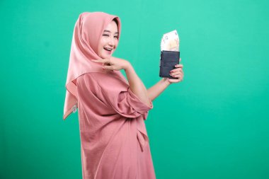 Tesettürlü genç bir kadın bir cüzdan dolusu Avro banknotunu sunarak, yeşil bir arka planda izole mali refah ve yatırım fırsatları öneriyor.