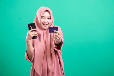 Cep telefonu ve ödeme kartı sunan neşeli tesettürlü kadın, yeşil arka planda izole edilmiş, uygun cep telefonu ödemeleri ve finansal teknolojileri tasvir ediyor..