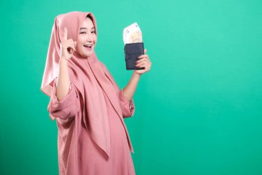 Başörtüsü içinde kendinden emin bir Müslüman kadın avro ve cüzdanını gösteriyor ve bu da başarılı bir finansal fikri simgeliyor. Yeşil arka planda izole edilmiş bu portre, finansal fikirleri destekleyen modern ve neşeli bir yatırımcıyı tasvir ediyor..