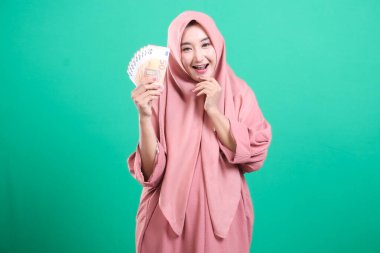 Mutlu bir ifadeye sahip neşeli bir kadın, tesettürlü ve avro banknotları tutan, yeşil bir arka planda izole edilmiş, bolluk taşıyan bir mali zafere sahip. 105 karakter