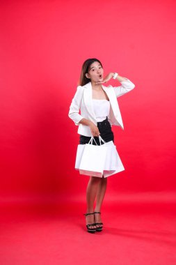 Beyaz önlüklü Endonezyalı kadın beyaz önlüklü, elleri çenesine kadar açılmış, beyaz kese kağıdı taşıyan, izole kırmızı arka planda duran. şirket içeriği, iş, işlem, alışveriş