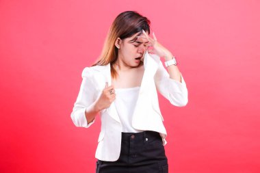 Beyaz ceketli ve siyah pantolonlu bir kadın kafasına ve göğsüne dokunuyor, aşağıya bakıyor, kırmızı arka planda soyutlanmış.