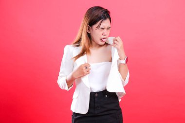 İğrenmiş bir yüzü olan genç bir kadın, beyaz bluz, siyah etek ve beyaz ceket giyiyor, üstünü tutuyor ve dilini dışarı çıkarıyor, kırmızı bir arka planda izole edilmiş.