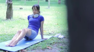 Genç bir kadın, huzurlu bir parkta paspasın üzerinde zarif bir yoga pozu verirken görülüyor. Atletik giyinmiş ve boynunda kulaklık var.,