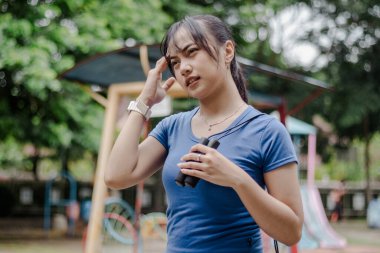 Genç bir kadın elinde halat tutuyor. Yorgun görünüyor. Egzersiz yaptıktan sonra baş ağrısı çekiyor. Sabahleyin parkta..