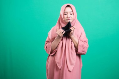 Başörtüsü içinde üzgün bir Müslüman kadın, mali kriz veya yoksulluğu temsil eden boş bir cüzdan taşıyor. Yeşil bir arka planda izole edilmiş bu görüntü ekonomik zorluk ve güvensizlik hissini yakalar..