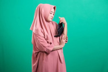 Başörtüsü takan düşünceli bir kadın, bir ayakkabıyı incelikle inceler, stil ve geleneklerini gösterir, yeşil bir arkaplanda izole edilmiş, çarpıcı ve çekici bir stüdyo portresi yaratır..