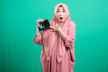 Başörtüsü takan şok olmuş Müslüman bir kadın yeşil arka planda izole edilmiş mali sıkıntıyı ve ekonomik sıkıntıyı simgeleyen boş bir cüzdan taşıyor. Borcunu ve birikimlerini dert ediyor. Bu resim kişisel finans krizini anlatıyor..