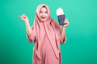 Zevkli Müslüman bir kadın tesettür takıyor, elinde avro banknotlarıyla bir cüzdan tutuyor ve yeşil arka planda izole edilmiş bir şeyi işaret ediyor..