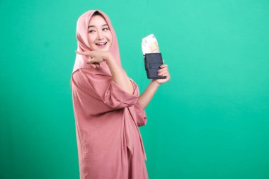 Tesettürlü neşeli bir kadının portresi yeşil bir arka planda izole edilmiş, avro banknotlarıyla dolu bir cüzdanı işaret ediyor ve tutuyor..