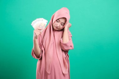 Avrupalı banknotların bir hayranını elinde tutan düşünceli Müslüman kadın, yeşil bir arka planda izole edilmiş bir yansıma anında gelecekteki mali kararları düşünüyor..