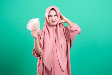 Başörtüsü içindeki bir kadın mali stres yaşar, elinde para varken endişeli görünür, yeşil bir zeminde izole edilmiş, maddi sıkıntılar ve potansiyel zorluklar gösterir..