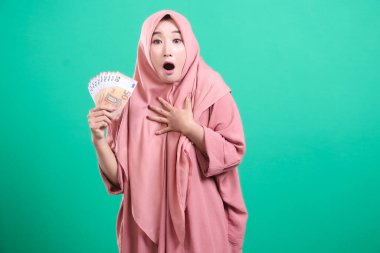 Şok olmuş Müslüman kadın elinde avro banknotlarıyla yeşil bir arka planda izole edilmiş büyük bir sevinç ve heyecan gösteren bir ifadeyle tesettüre sarılmış.