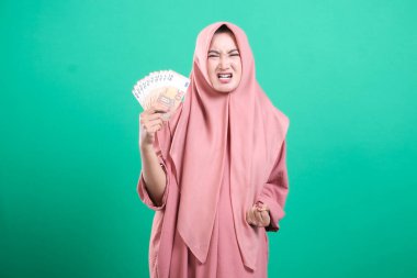 Kızgın Asyalı İslamcı kadın tesettüre girmiş ve nakit para gösteriyor. Ekonomik dengesizlik ve kişisel finans sorunları, yeşil bir arka planda izole.