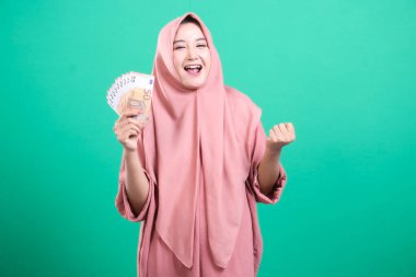 Avro banknotları tutan ve gülümseyen tesettürlü neşeli Müslüman kadın, mutluluğu ifade ediyor ve başarıyı kutluyor; yeşil bir arka planda izole edilmiş. Finansal güvenliği, kazancı ve kazanma anlarını somutlaştırıyor..