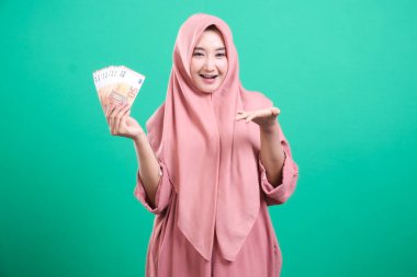 Avro banknotları tutan, elinde jest yapan, yeşil arka planda izole edilmiş neşeli bir ifadeyle gülümseyen Müslüman bir kadın..