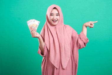 Gülümseyen neşeli Müslüman kadın elinde Avro banknotlarıyla sağ tarafı işaret ediyor. Yeşil arka planda izole edilmiş, finansal refah ve ekonomik fırsatları iletiyor..