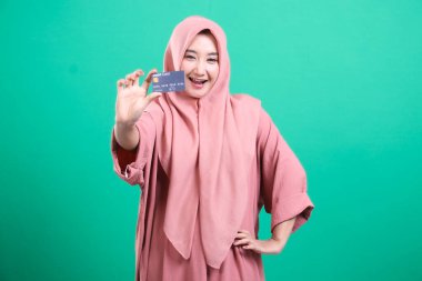 Kredi kartı modeli gösteren neşeli Müslüman bir kadın pozitiflik ve tüketici güveni yayıyor. Parlak gülüşü, yeşil bir arka planda izole edilmiş güveni ve finansal gücü aktarıyor..