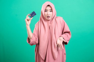 Endişeli bir ifadeye sahip genç bir Müslüman kadın kredi kartı gösteriyor, bu da yeşil arka planda mali sıkıntı veya alışveriş sorunu olduğunu gösteriyor. Şık, pembe bir elbise ve tesettüre bürünüyor..