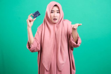 Kafası karışık bir ifadeyle kredi kartı tutan kafası karışmış Müslüman bir kadın, muhtemelen maddi zorluklarla boğuşuyor. Yeşil arkaplanda izole edilmiş.