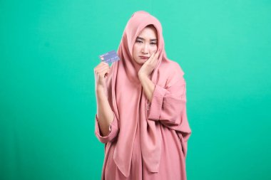Depresyondaki Müslüman kadının elinde kredi kartı var. Finansal stres ve endişeyi tasvir ediyor. Yeşil arka planda izole edilmiş, çağdaş toplumda borç yükünü simgeliyor..