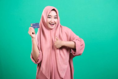 Başörtüsü içinde gülümseyen Müslüman bir kadının portresi, bir kredi kartı tutuyor ve başparmağını kaldırıyor, parasız ödemelerin başarısını temsil ediyor, yeşil bir arka planda izole edilmiş. Modern finansal hizmetleri ve teknolojiyi olumlu bir şekilde kucaklamak.