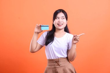 Mutlu genç bir kadın bir kredi kartı tutuyor ve kenarı işaret ediyor. Bu da turuncu arka planda izole edilmiş finansal hizmetlerle ilgili büyük bir fırsat ya da özel bir teklif sunuyor.