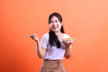 Neşeli genç bir kadın elinde bir tabak lezzetli şehriye tutarken sol tarafı göstererek turuncu arka planda izole edilmiş büyük bir gülümsemeyle.